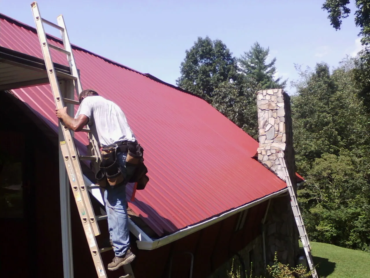 Roofer on a ladder beside a red metal roof and stone chimney — Soffit & Fascia Repair by Roofing Brothers in Eagle Point